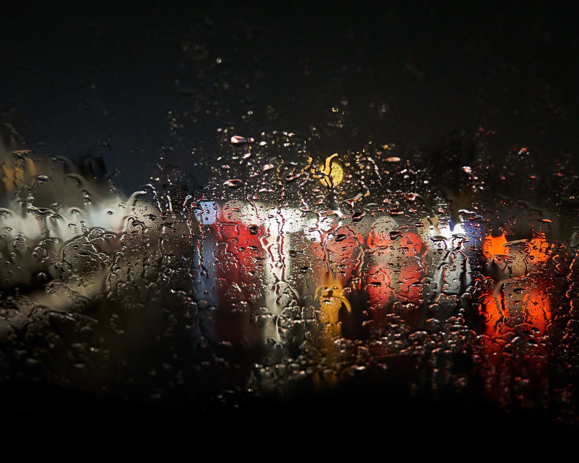 Rain-streaked windshield at night, red and amber headlights and taillights smeared into long halogen streaks through the water.