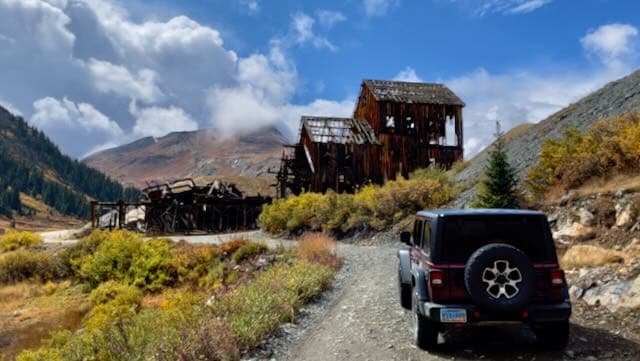An off-road vehicle beside a mountain landscape and a weathered structure under changing clouds.