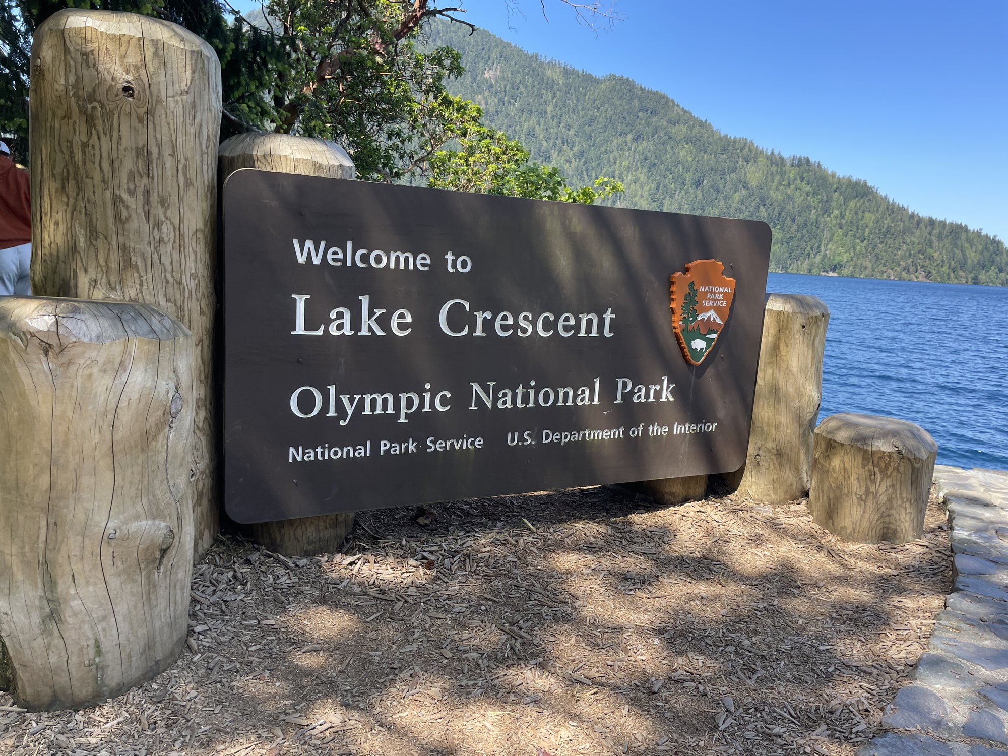 The Lake Crescent and Olympic National Park welcome sign beside blue water.